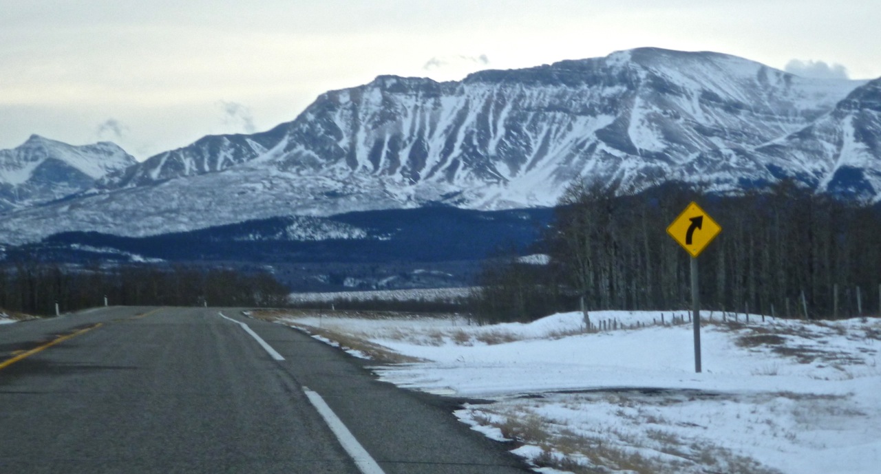 Straße, im Hintergrund Berge, rechts ein Kurvenschild